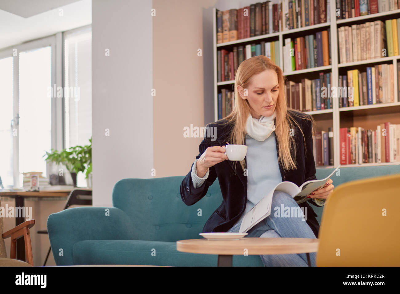 one woman, one person 40 years old, holding coffee cup, reading magazine, in library, book store, book shop, sitting in sofa. Shelf full of books behi Stock Photo