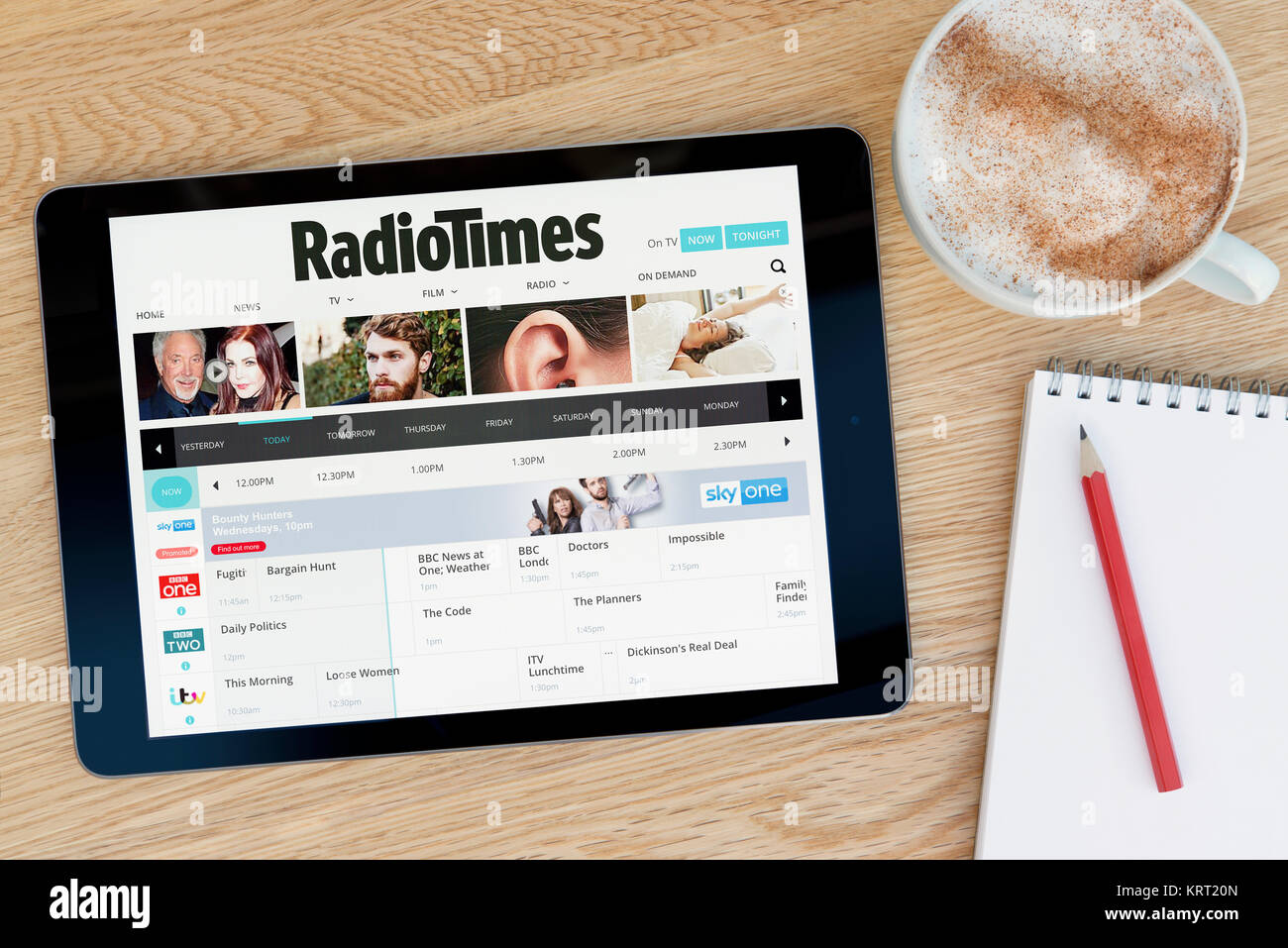 The Radio Times website on an iPad tablet device which rests on a wooden table beside a notepad and pencil and a cup of coffee (Editorial only) Stock Photo