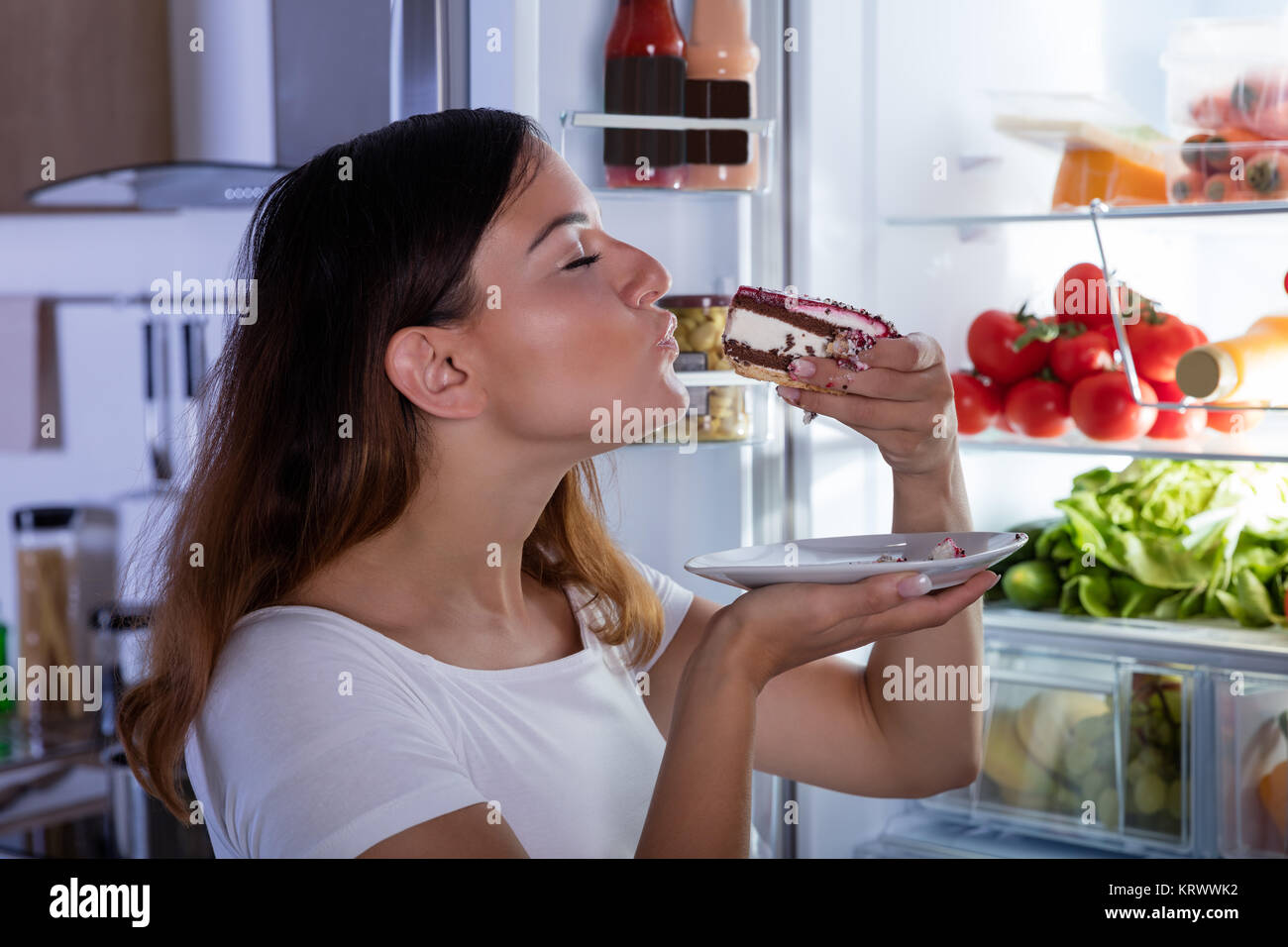 Woman Eating Cake In Front Of Refrigerator Stock Photo