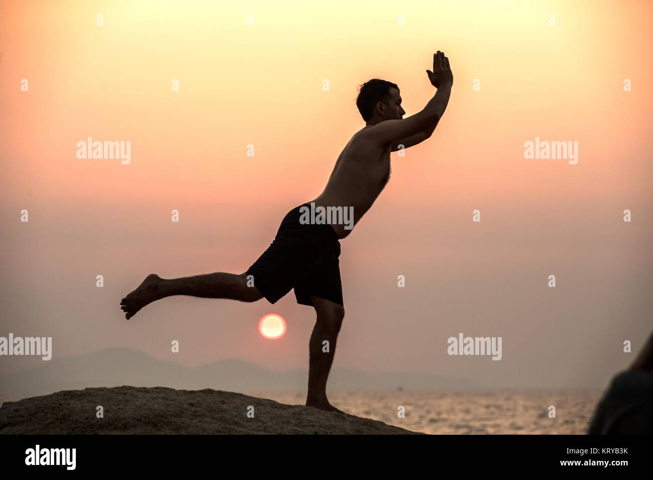Positive male holding hand up and expressing gladness while standing on stone in Sunset. Stock Photo