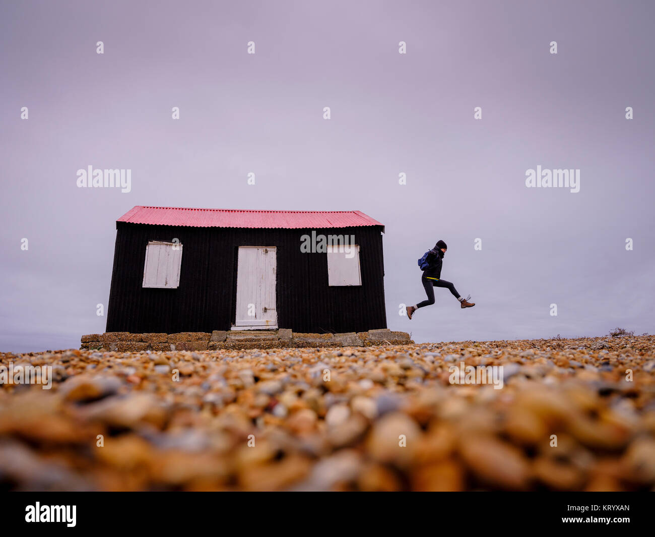 A woman leaps from the side of a small black and red hut on the riverbank in Rye Harbour Nature Reserve, East Sussex UK Stock Photo
