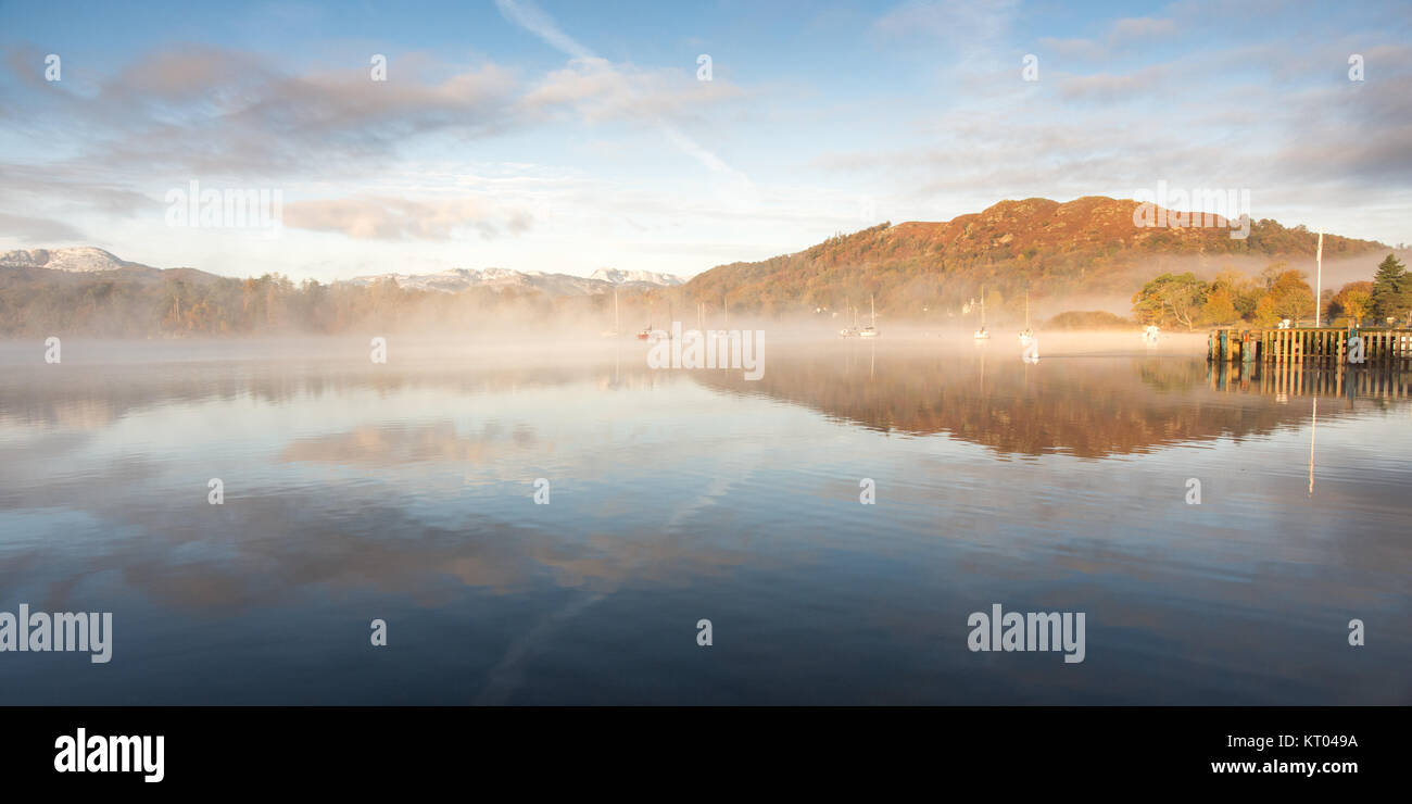 Mist rises from Windermere lake, beside woodland trees in autumn colours, at Ambleside in England's Lake District National Park. Stock Photo