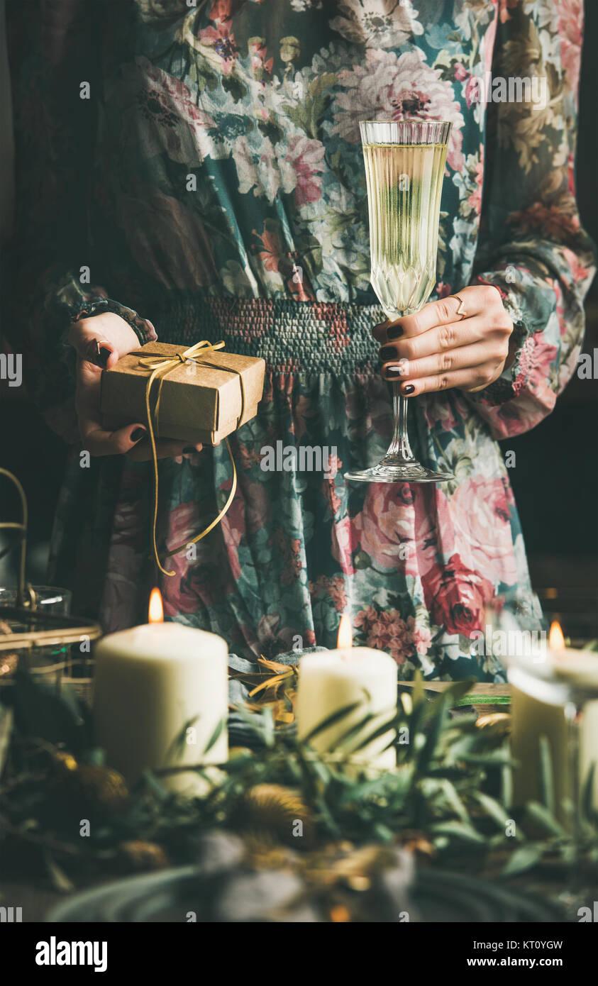 Woman in festive dress holding glass of champaigne and gift box in her hands at holiday decorated table during Christmas or New Year celebration party Stock Photo