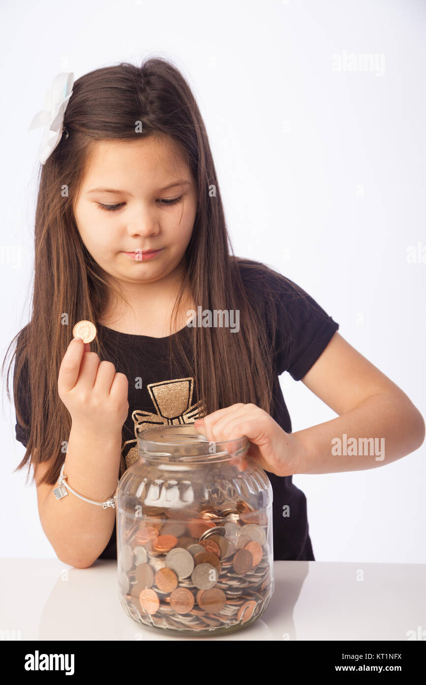 Seven year old girl holding  a penny coin and looking into a jar full of coins. Stock Photo