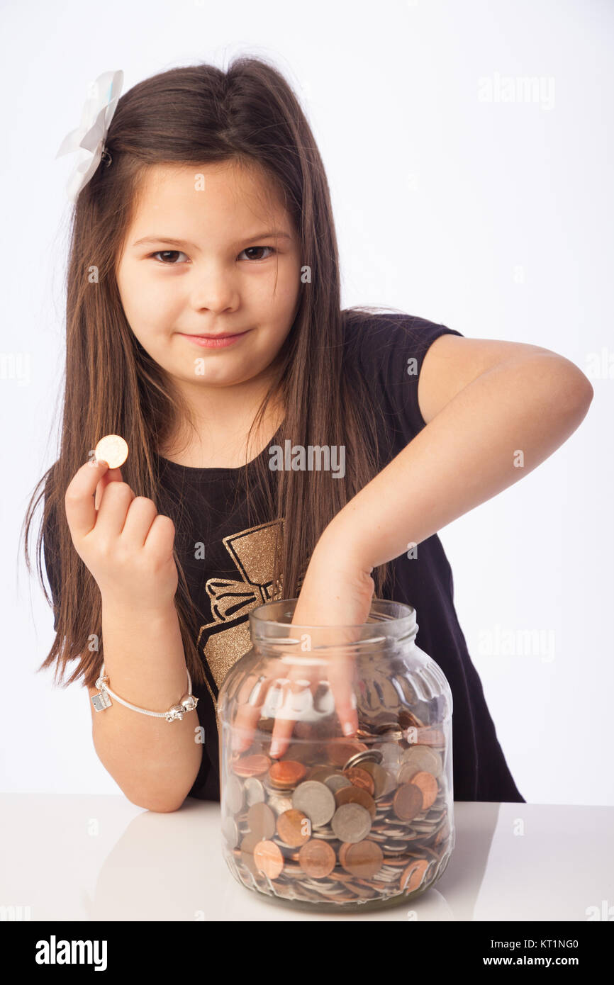 A seven year old girl holding a penny coin in one hand and with the other grabbing coins inside a glass jar. Stock Photo