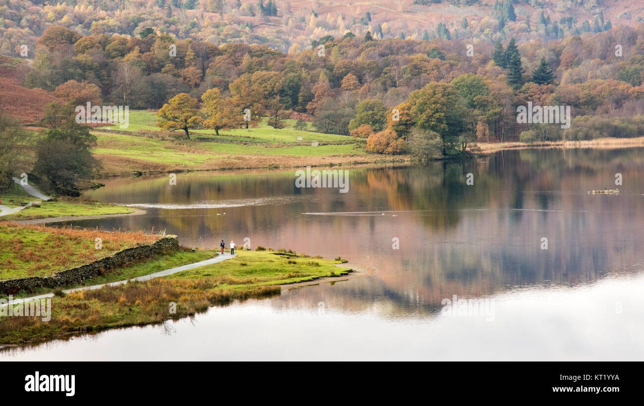 Hikers walk alongside Rydal Water lake beside trees displaying autumn colours in England's Lake District National Park. Stock Photo