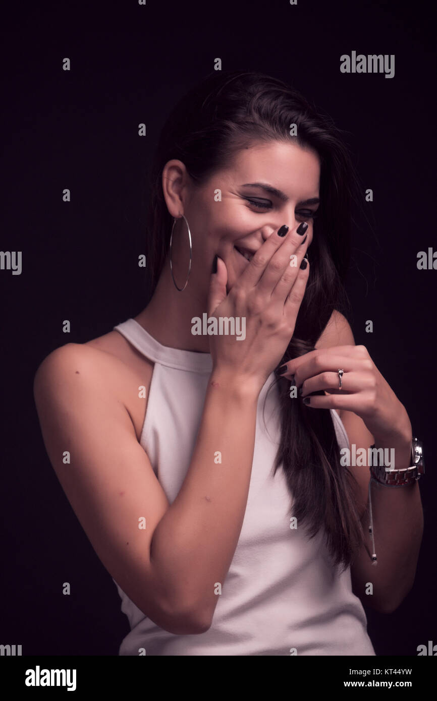 one young beautiful woman portrait, sincere candid giggling, covering her mouth with hand. black background, white dress, studio. upper body shot. Stock Photo