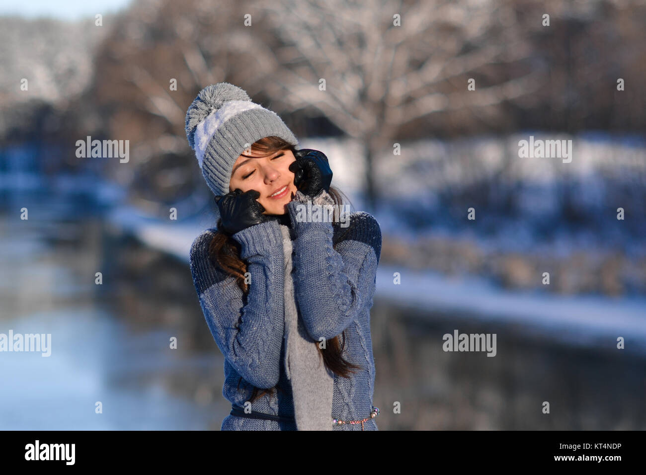 Picture of closed eyes sunny smiling beautiful cute girl enjoying a winter nature on winter snowy forest background. Portrait of beautiful girl in warm hat with balabon, sweater, scarf. Stock Photo