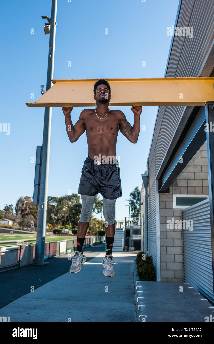 Muscular Jamaican athlete performing pullup exercise with hands apart on steel beam at stadium