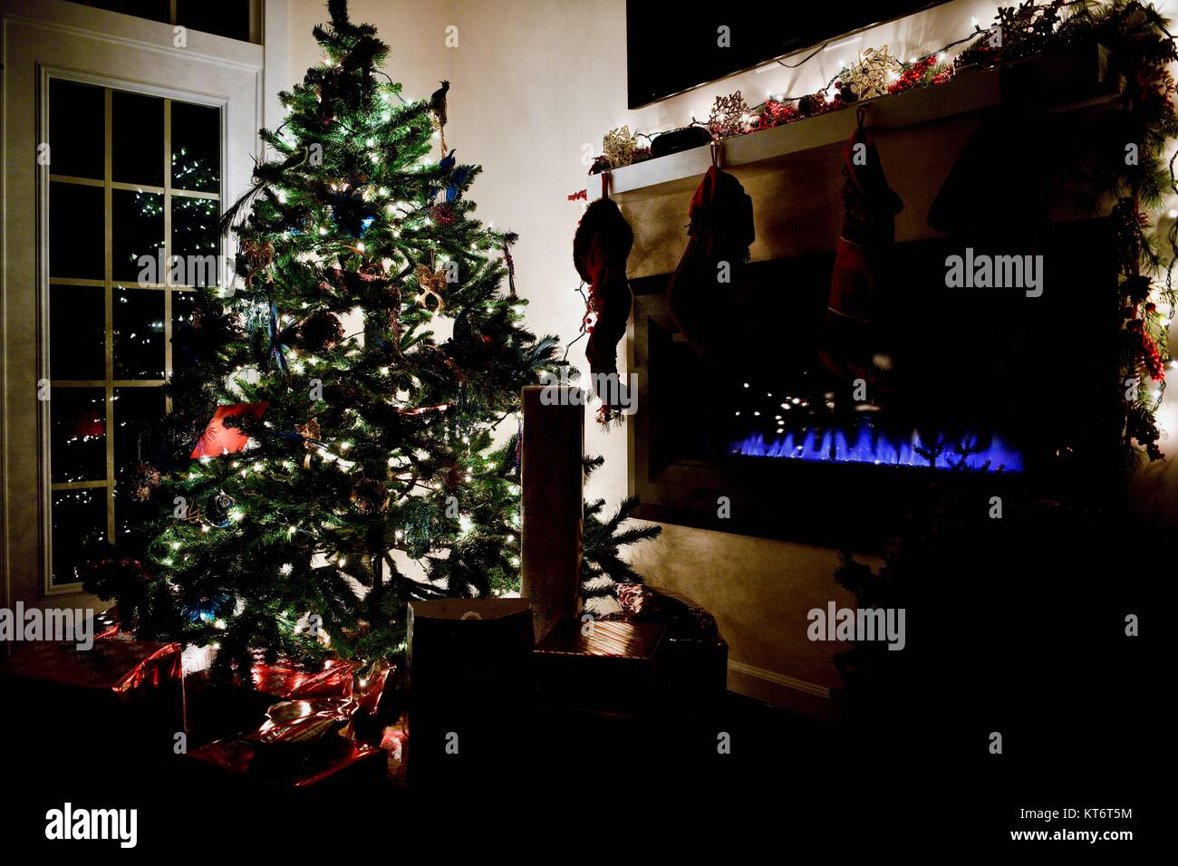 Christmas tree full of ornaments and lights with wrapped presents underneath next to a cozy gas