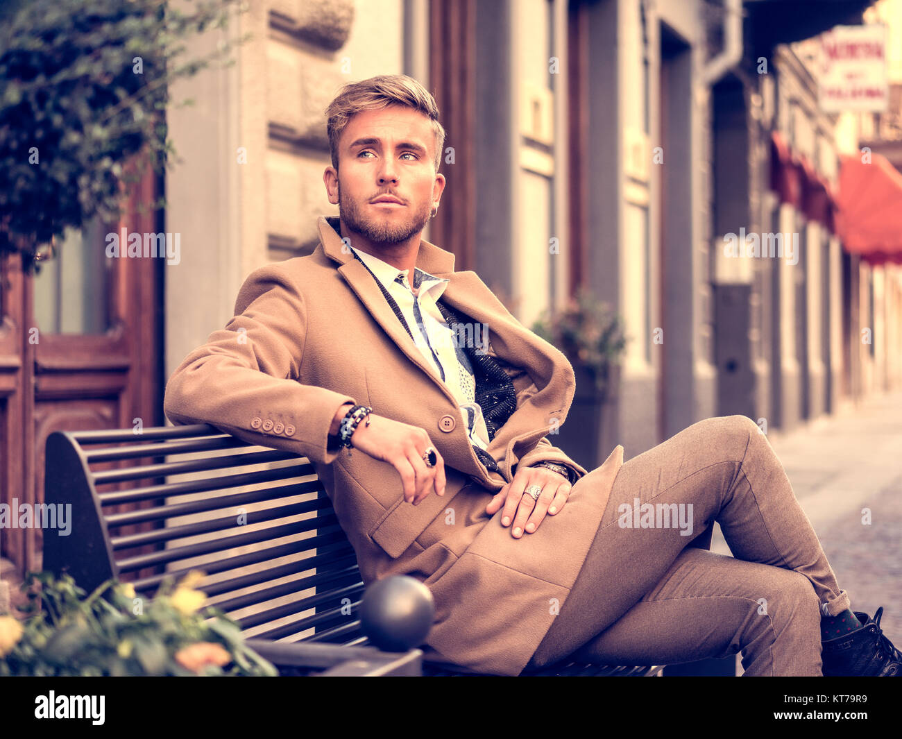 handsome man wearing elegant coat sitting on bench at street and posing KT79R9