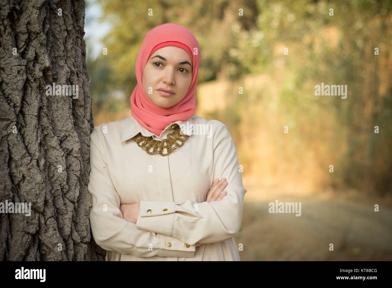 Beautiful Muslim woman wearing hijab leaning against a tree arms folded looking away Stock Photo