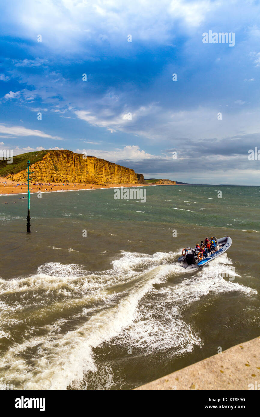 An inflatable rib full of tourists speeds out of the entrance to West Bay harbour in front of East Cliff on the Jurassic Coast, Dorset, England, UK Stock Photo