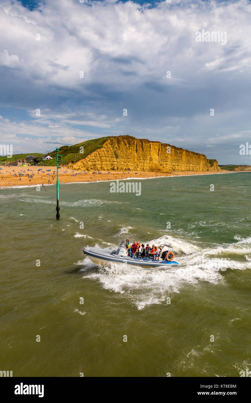 An inflatable rib full of tourists approaches the entrance to West Bay harbour in front of East Cliff on the Jurassic Coast, Dorset, England, UK Stock Photo