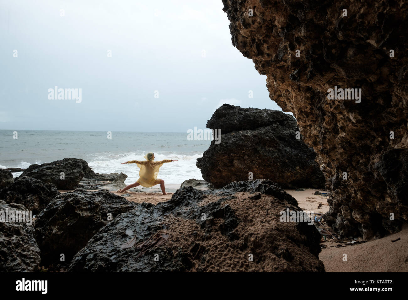 Woman in rain coat doing stand yoga asana on ocean beach during storm. Concept of yoga travelling on Bali Stock Photo