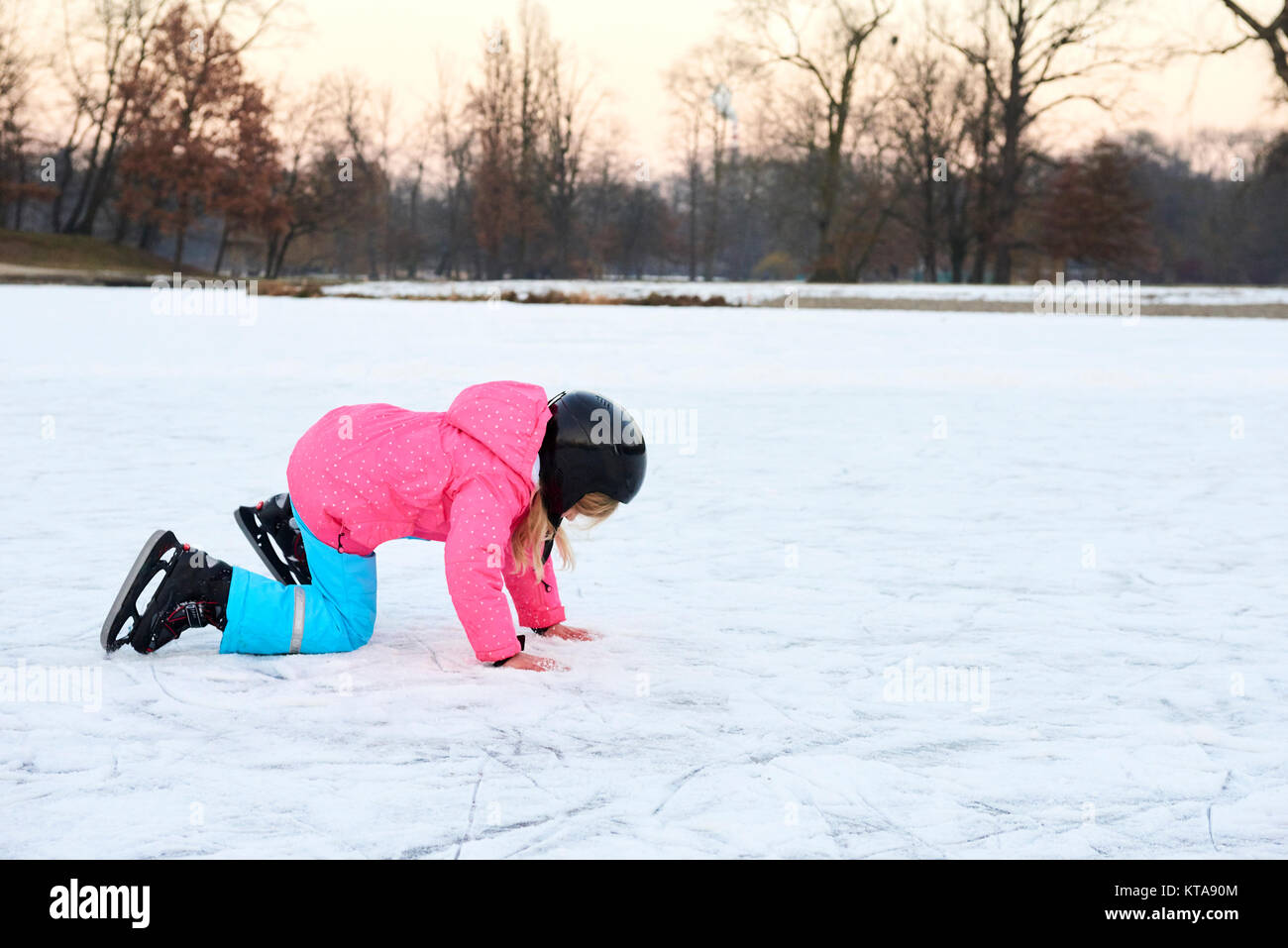 Child girl falling down on ice in snowy park during winter holidays. Wearing safety helmet. Winter children activities. Stock Photo