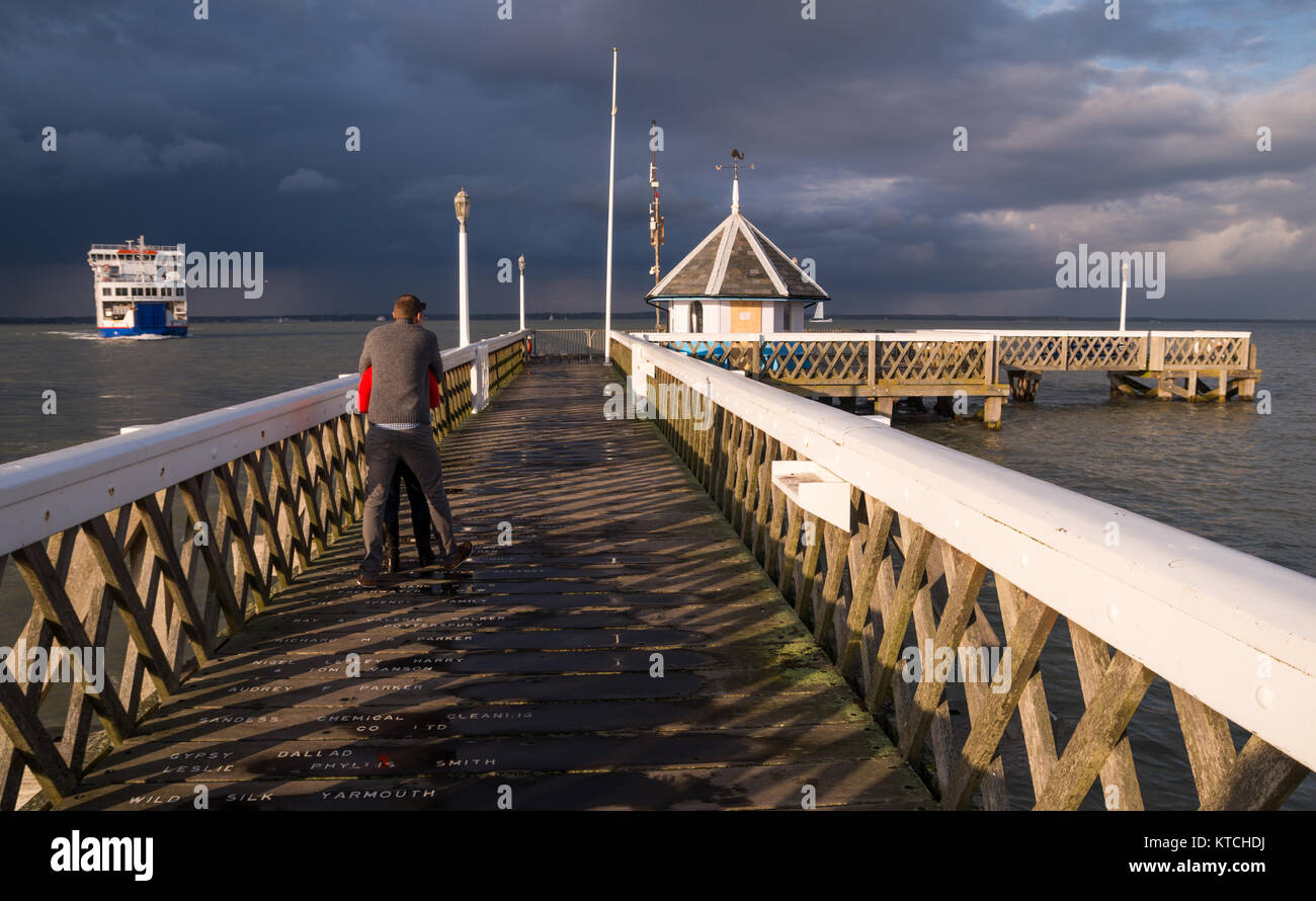 A scene on Yarmouth pier with a couple watching the approaching ferry and heavy grey clouds rolling in from the mainland in warm evening light. Stock Photo