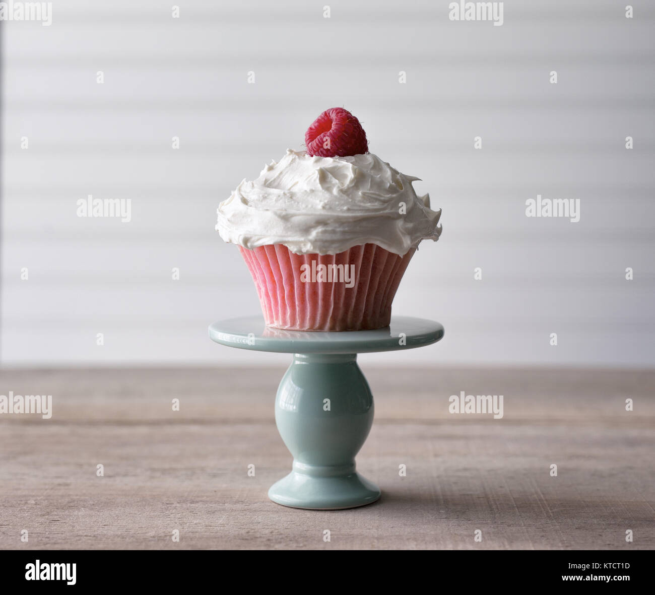 Pink cupcake with fresh raspberry garnish being served from a mini cake plate. Against a white background. Copy space. Stock Photo