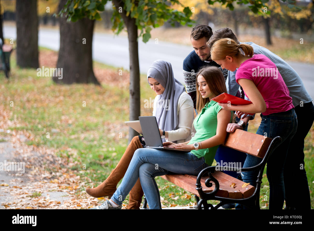 Group Of Young People Using Laptop And Tablet On A Park Bench