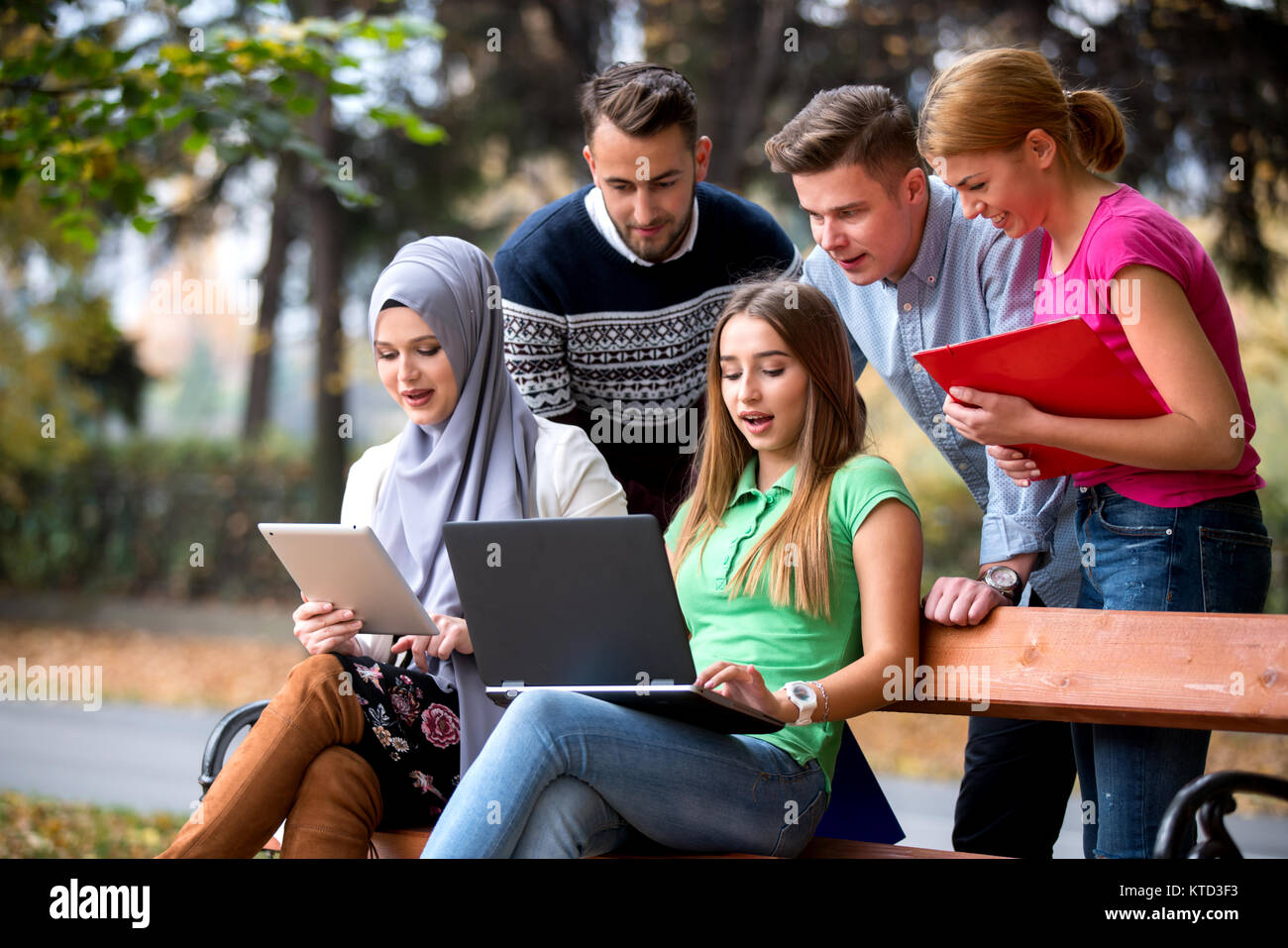 Group Of Young People Using Laptop And Tablet On A Park Bench