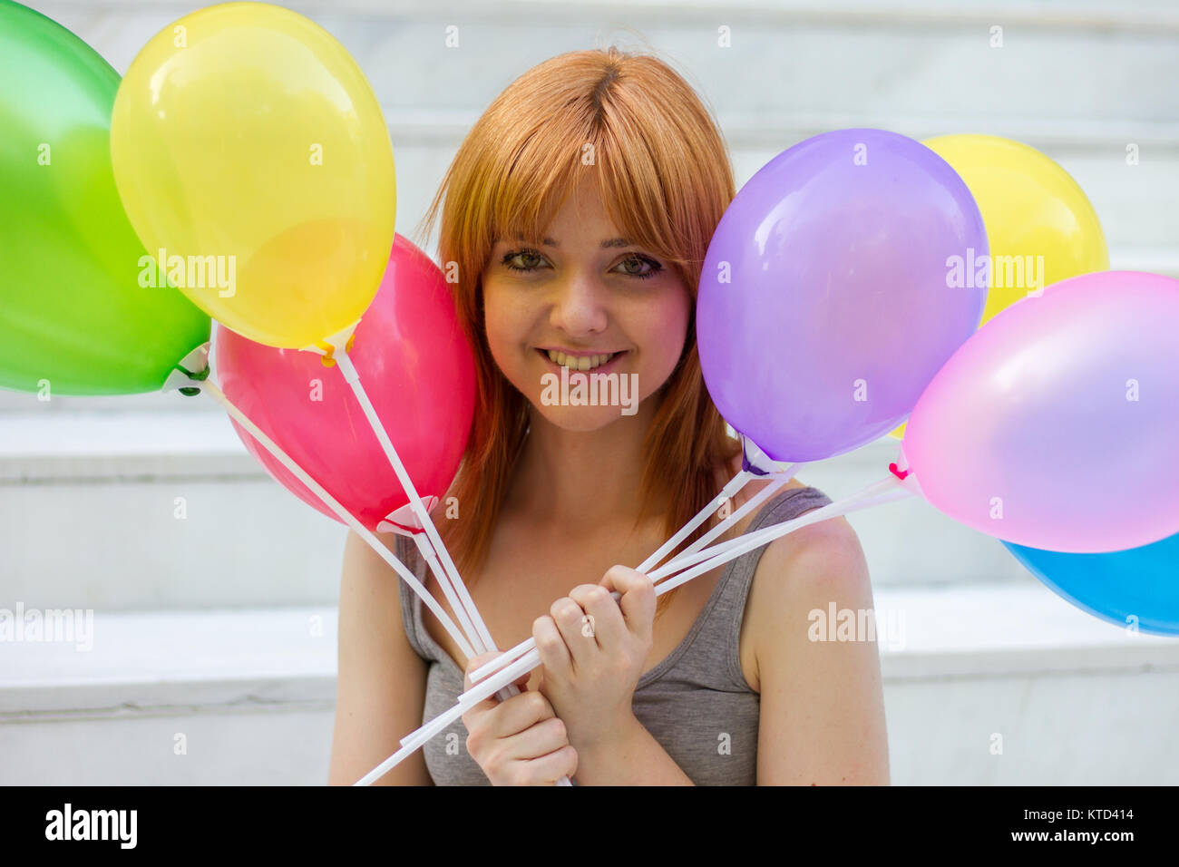 Portrait of a white girl with orange hair sitting on a white stairs holding colorful balloons with their hands. Stock Photo