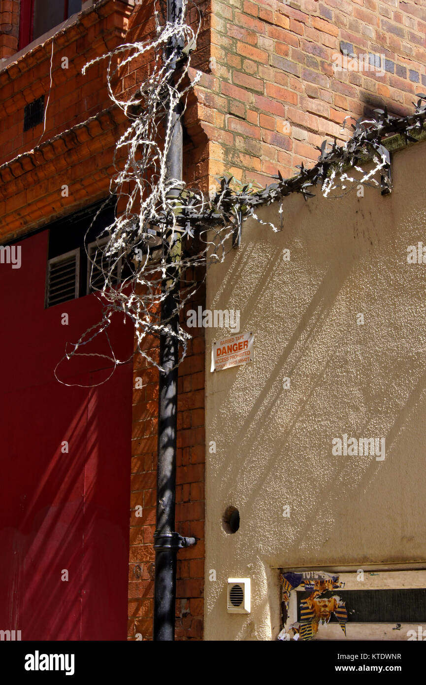 Razor wire looped round a black drainpipe, next to a red door, red brick wall and white pebbledash wall, with air vents and a red warning sign Stock Photo