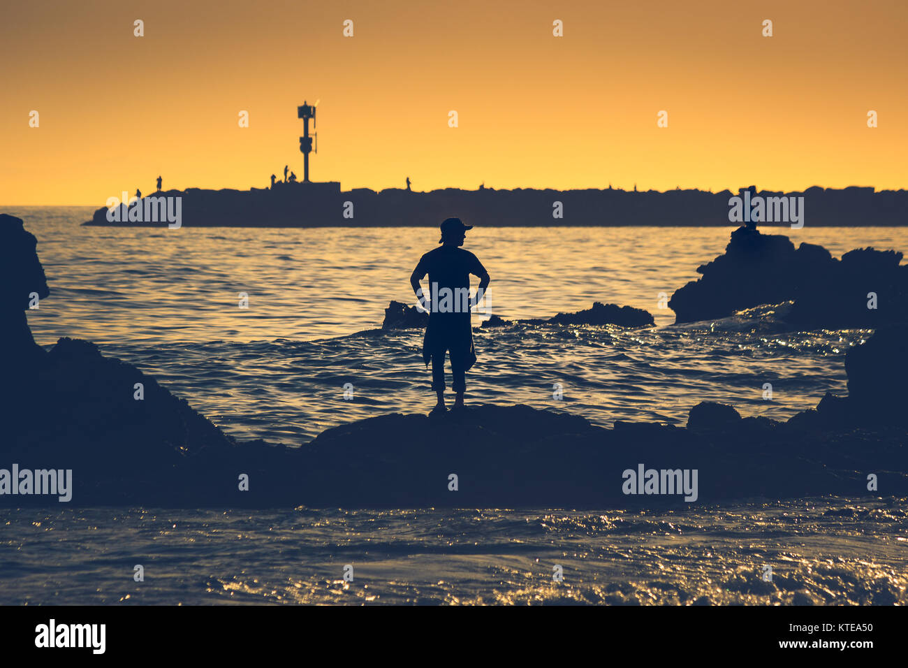 Silhouette of man posed on a rock formation at sunset from Little Corona Beach, Corona Del Mar, California with people fishing from a jetty in the bac Stock Photo
