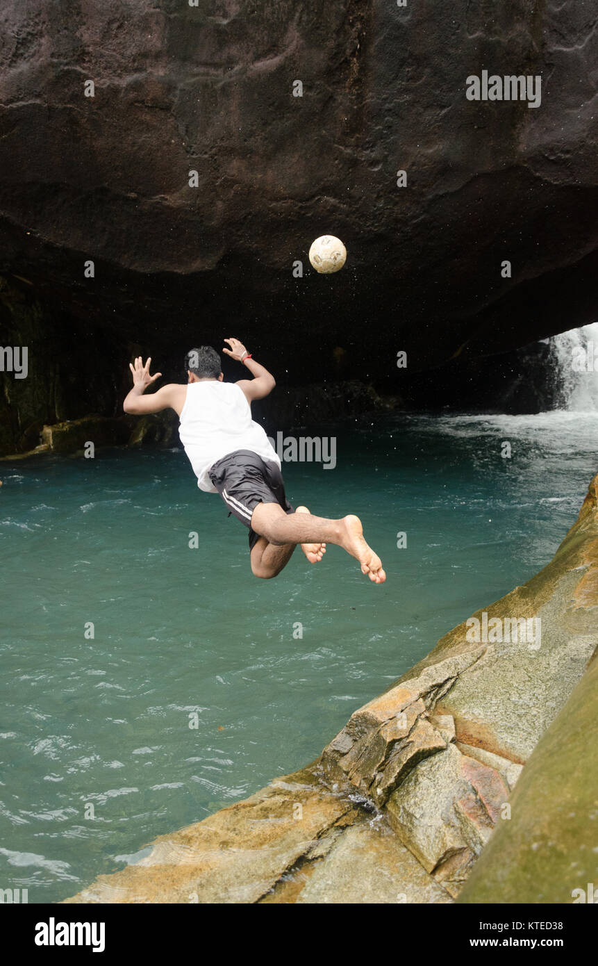 Back view of a boy diving off cliff, throwing away a ball, falling unprofessionally into blue waters at Nagarmadi Water Falls, Chendia, Karnataka Stock Photo