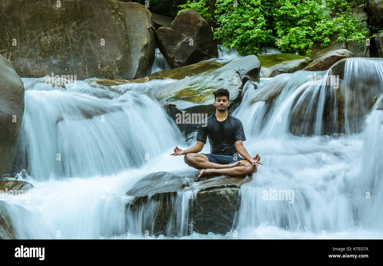 Young adult boy sitting on a rock midst smooth splashing and flowing water, meditating at a portion of Nagarmadi Falls in Chendia, Karnataka, India Stock Photo