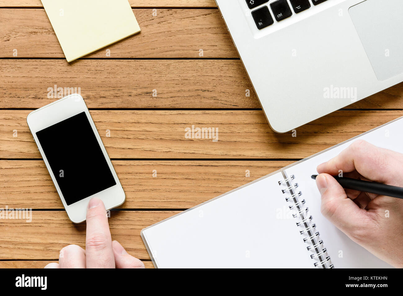 Overhead view of a neat wooden desktop with male hand writing on a notebook and touching a smartphone, laptop and notepad. Stock Photo