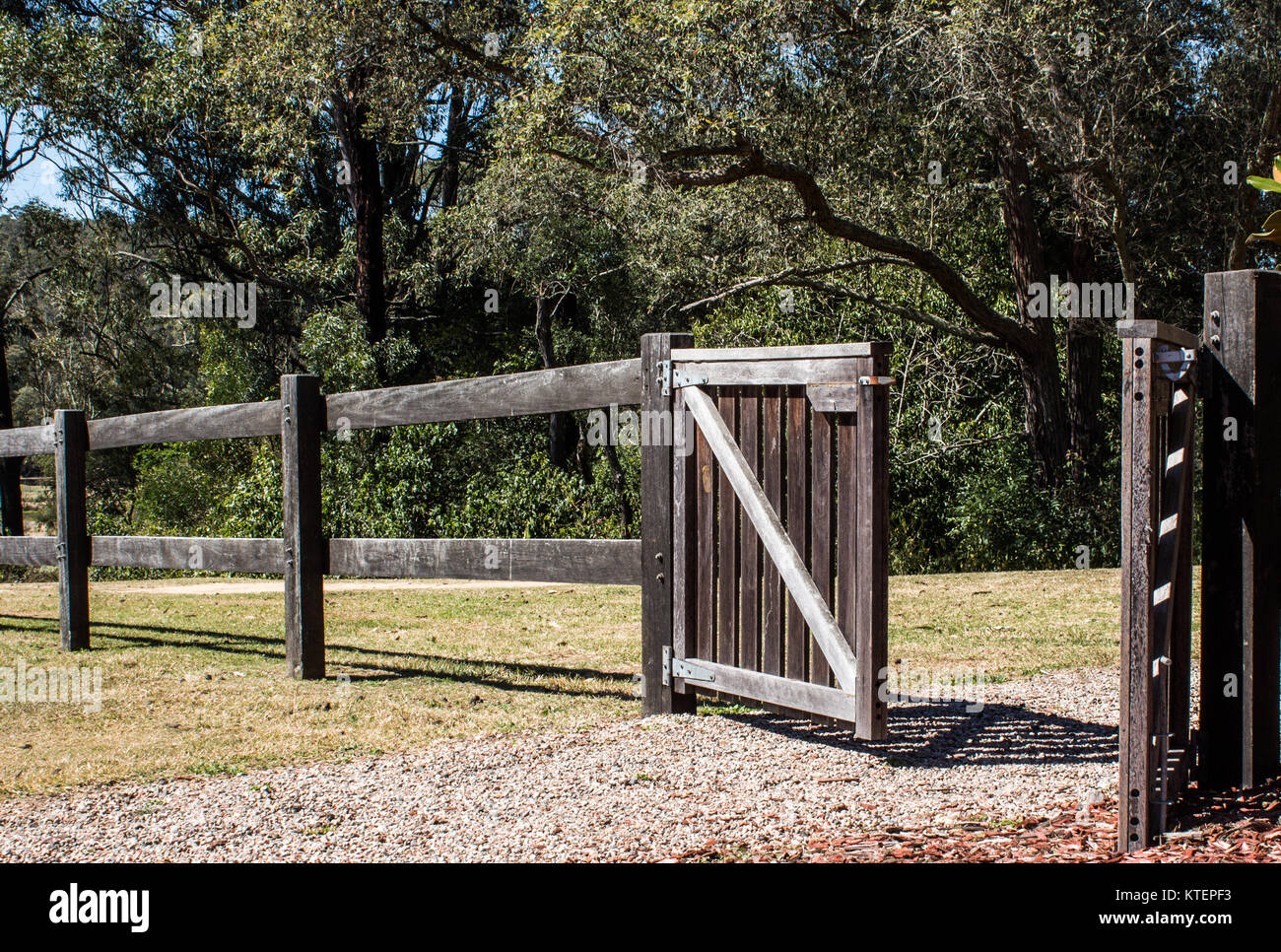 Wooden rural fence with open gate in sunshine with trees in background Stock Photo