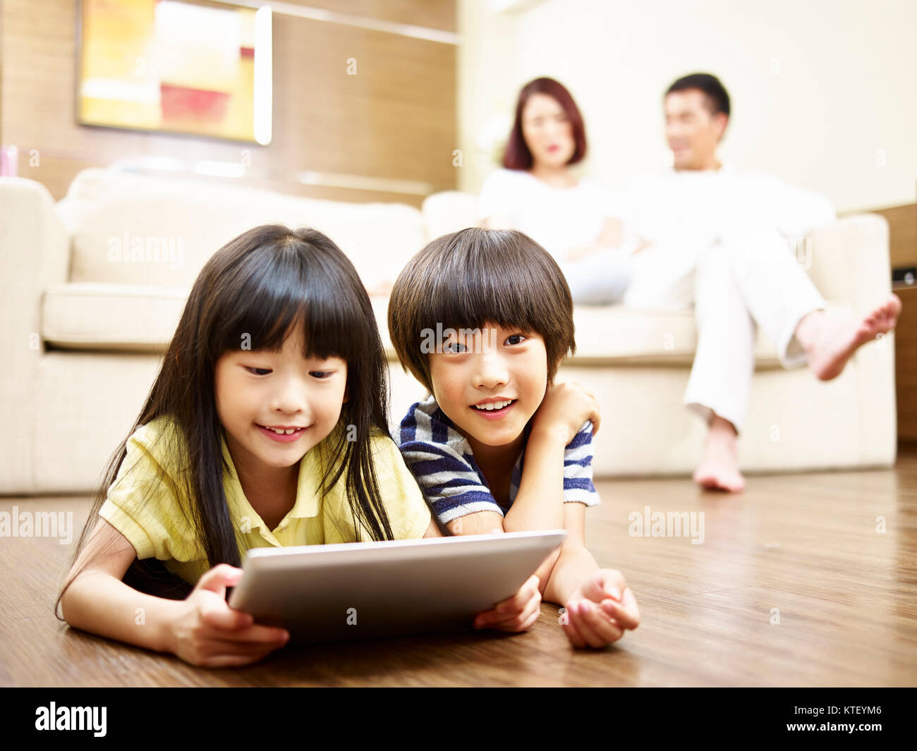 two asian children lying on floor playing video game using digital tablet while parents watching in the background. Stock Photo