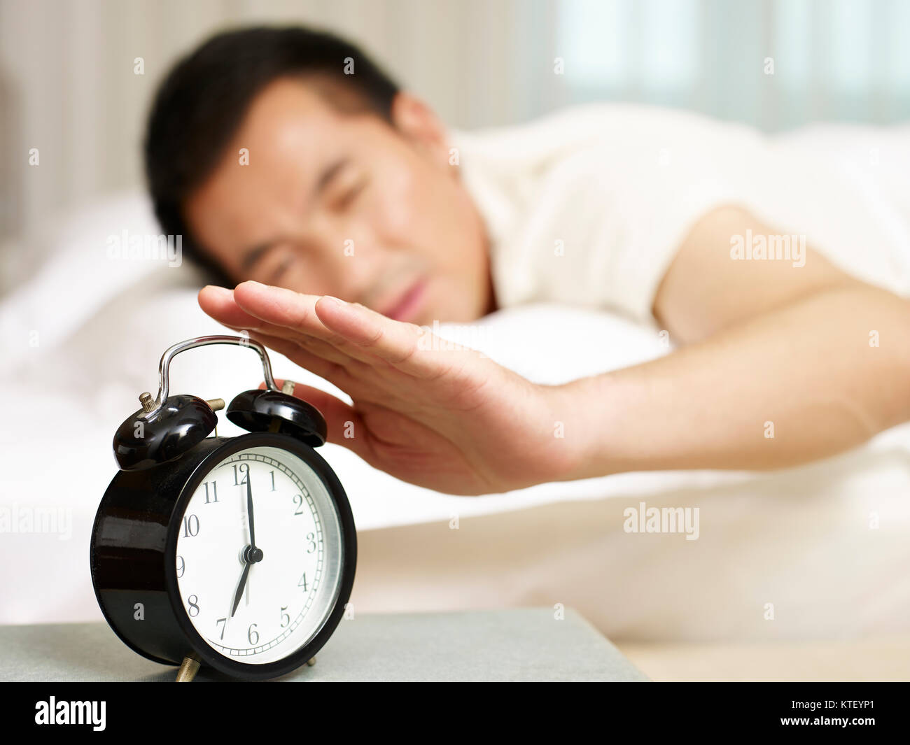 asian man lying in bed trying to stop ringing alarm clock, focus on the clock. Stock Photo