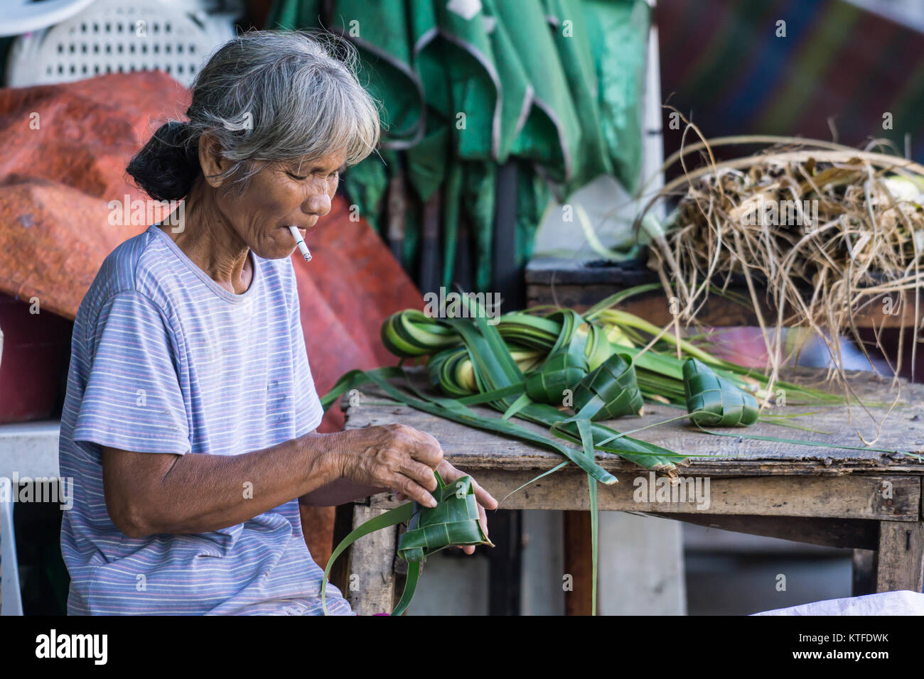 An elderly woman making Puso,a woven triangular basket of young coconut leaves in which rice is cooked. Stock Photo