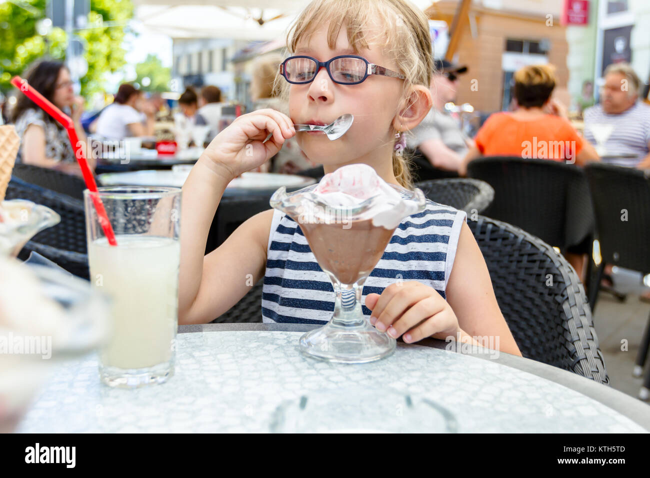 Little cute girl with glasses and carefree face expression is eating ice cream at table in pastry shop in summertime. Stock Photo