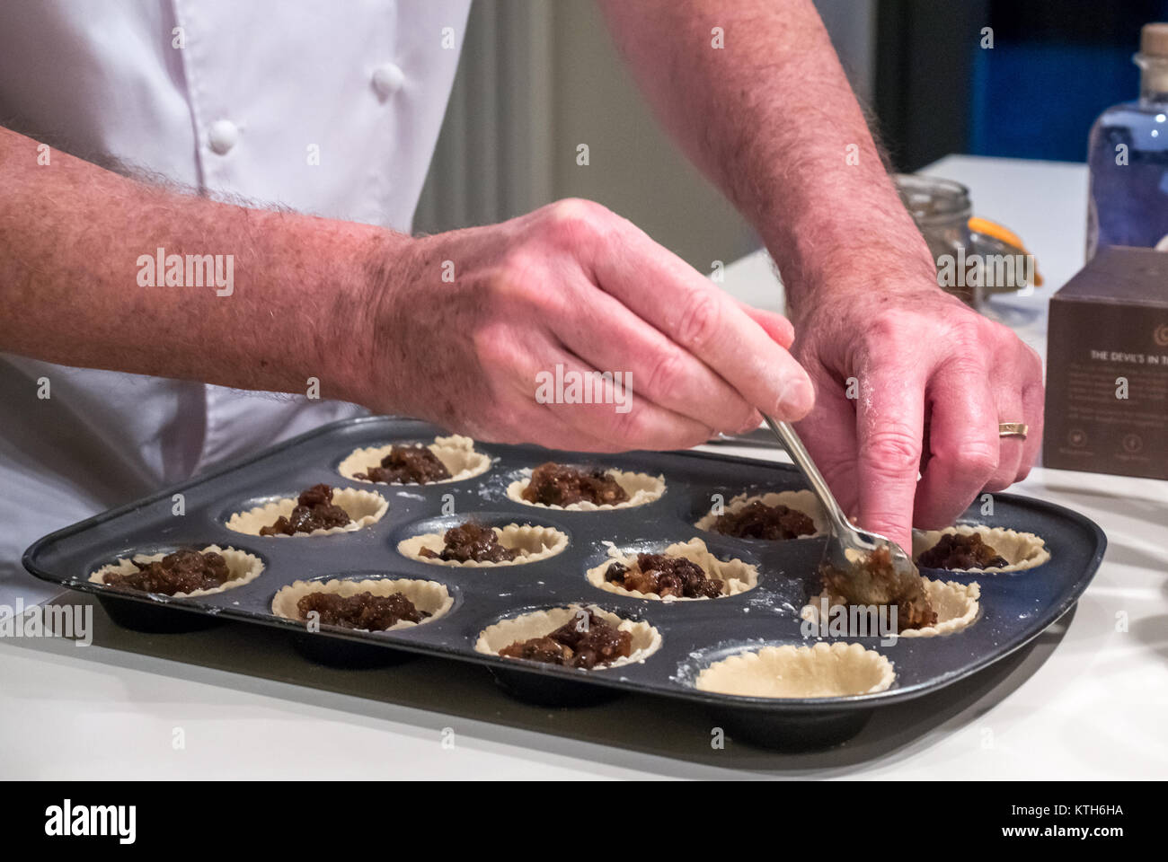 Close up of older man in chef whites filling mince pie cases with mincemeat ready for baking at Christmas time on a white kitchen counter Stock Photo