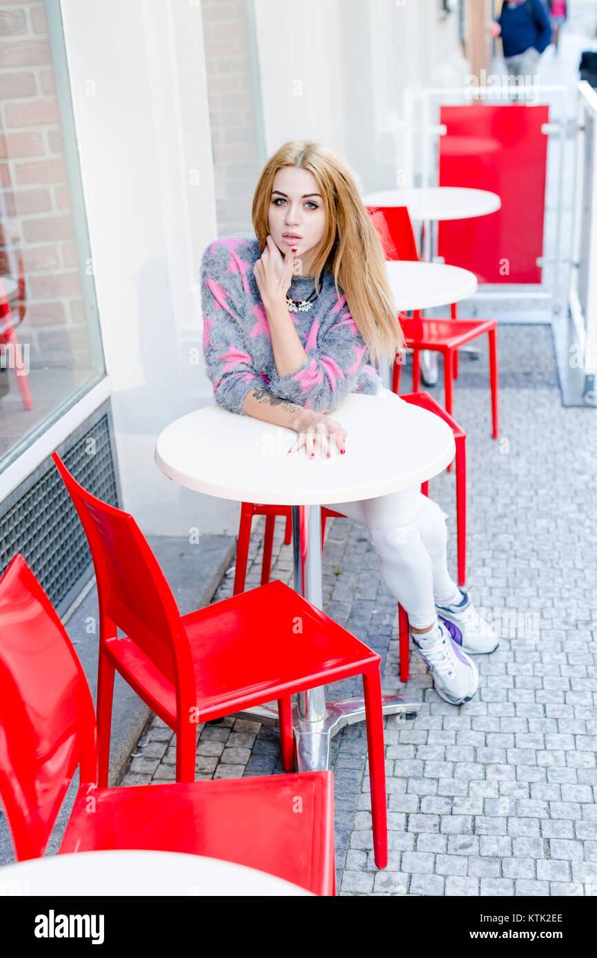 Portrait of a cute blonde girl sitting in a outdoor cafe with red chairs and empty table Stock Photo