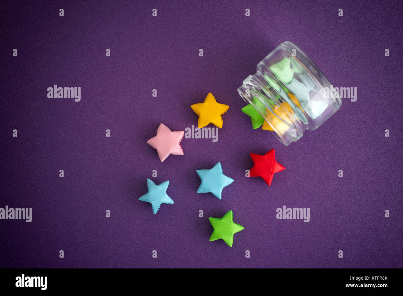 Colourful origami lucky stars spilling out of a jar. Purple background. Stock Photo