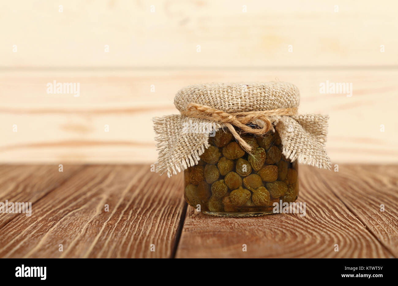 Close up of one small glass jar of pickled capers with canvas top decoration and twine on brown wooden table over white background, low angle side vie Stock Photo