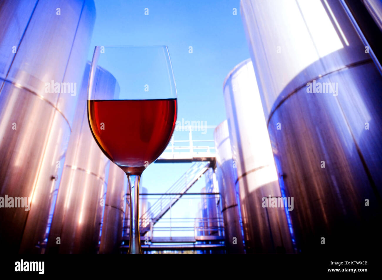 WINE MAKING CONCEPT CALIFORNIA Modern winemaking manufacture tasting concept, with glass of red wine in foreground and sunlit outdoor grape maturation tanks behind in perspective California USA Stock Photo