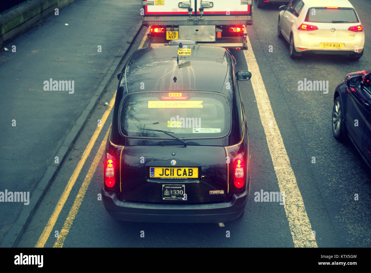 London black taxi cab on the road street between yellow lines with apt licence plate cab stuck in traffic behind  lorry truck in perspective red light Stock Photo