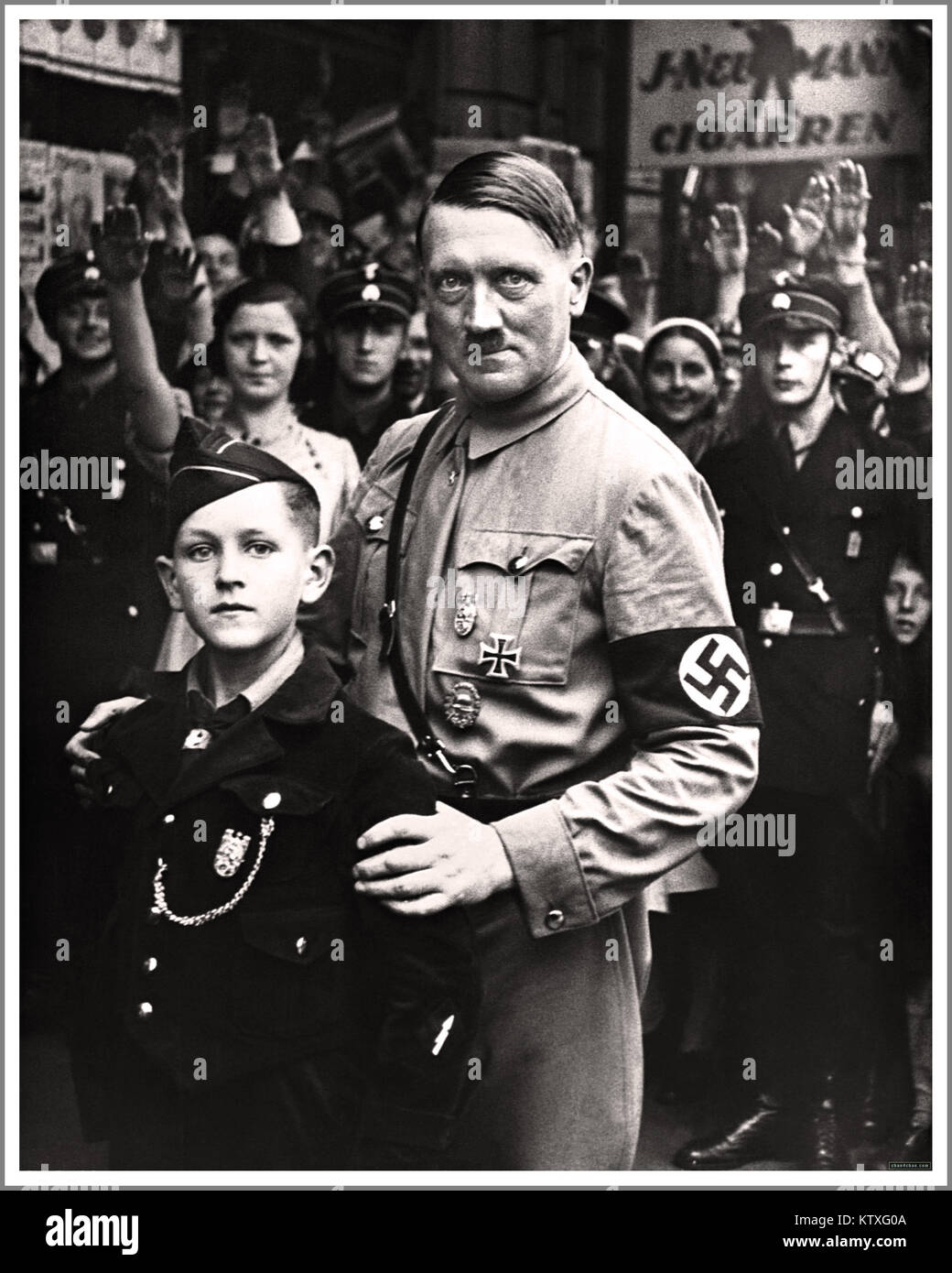 HITLER Nazi Germany Propaganda Image 1930’s Nazi dictator Adolf Hitler wearing NSDAP uniform and swastika armband in Berlin, posing with a chilling expression alongside a 10-12 years young boy member of the Hitler Nazi Youth Hitler-Jugend organisation. Crowd behind saluting Heil Hitler. Stock Photo