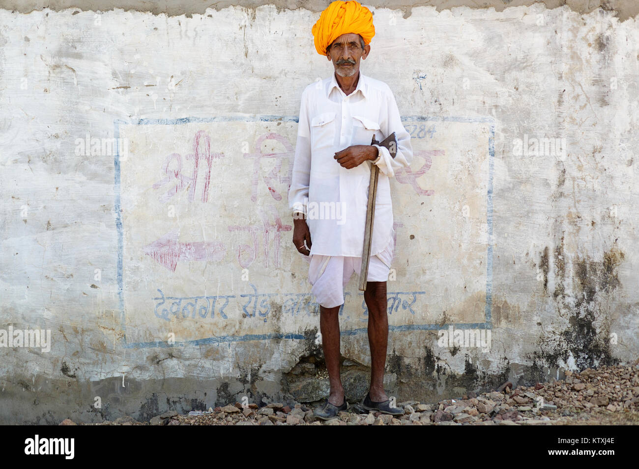Old indian man in white outfit wearing orange turban, standing in front of white wall in a village near Pushkar, Rajasthan, India. Stock Photo