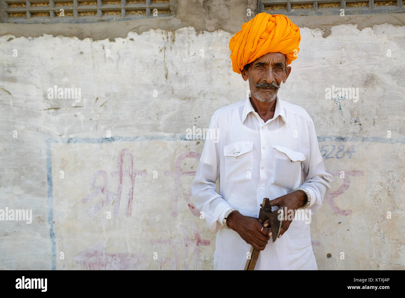 Old indian man in white outfit wearing orange turban, standing in front of white wall in a village near Pushkar, Rajasthan, India. Stock Photo