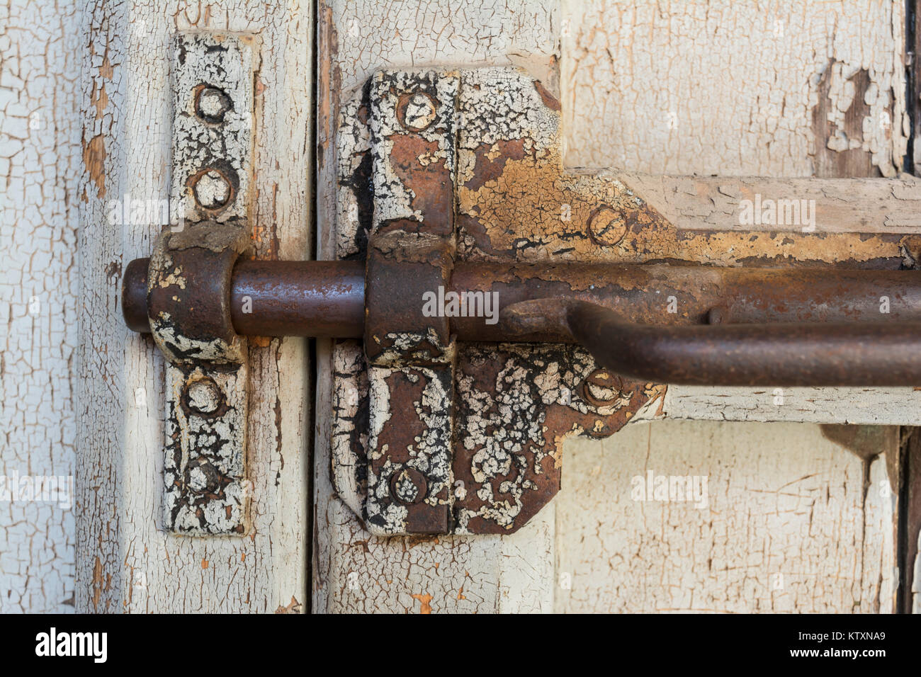 Very old, deteriorating gate latch bolt in a locked position, complete with rusting parts, and peeling white paint on the wooden door. Closely cropped a Stock Photo