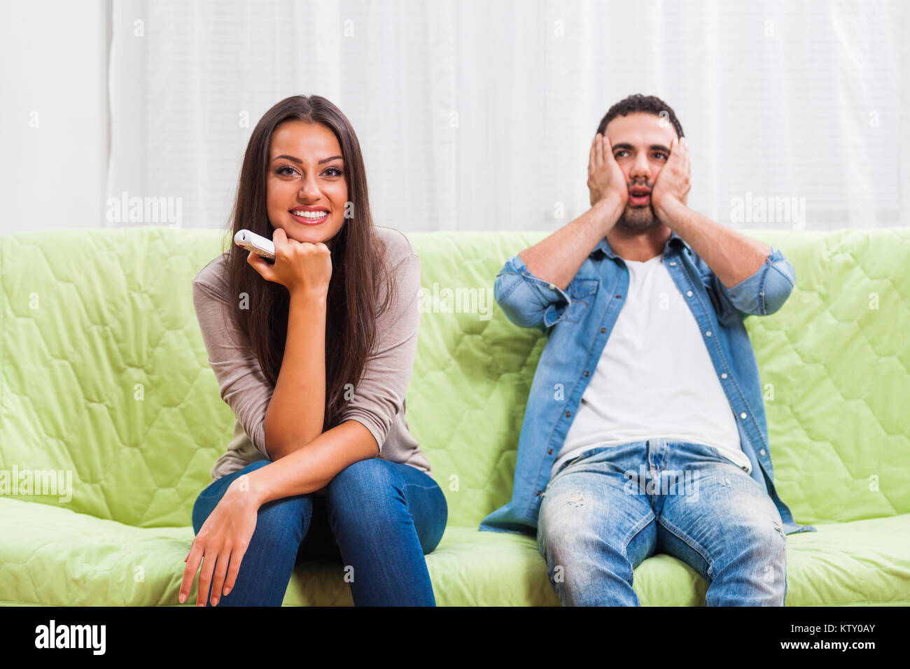 Young couple is sitting on sofa at home. Man is frustrated because he does not like what his woman is watching on TV. Stock Photo