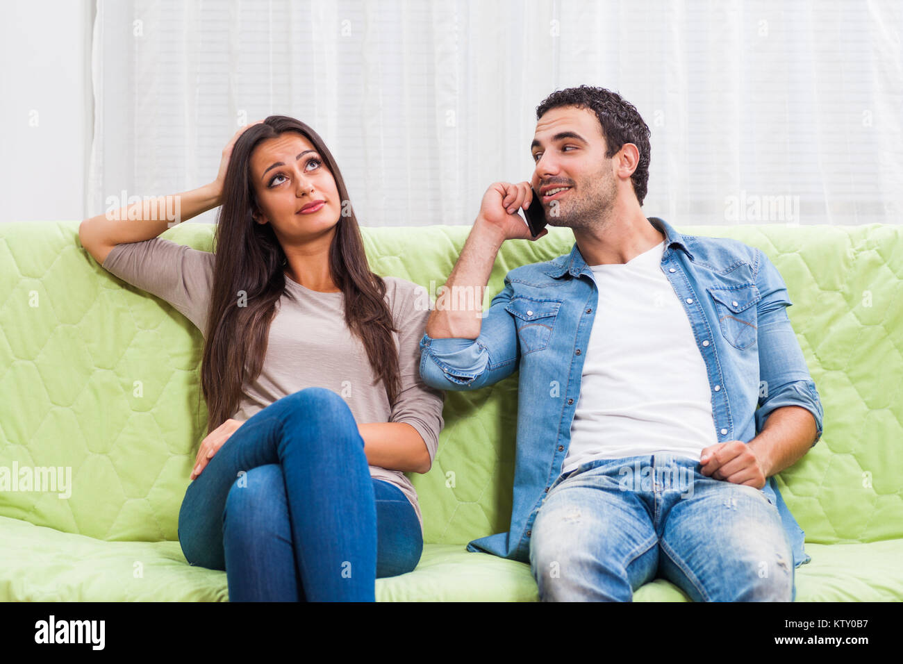 Young couple is sitting on sofa at home. Woman is getting bored while man is typing on phone. Stock Photo