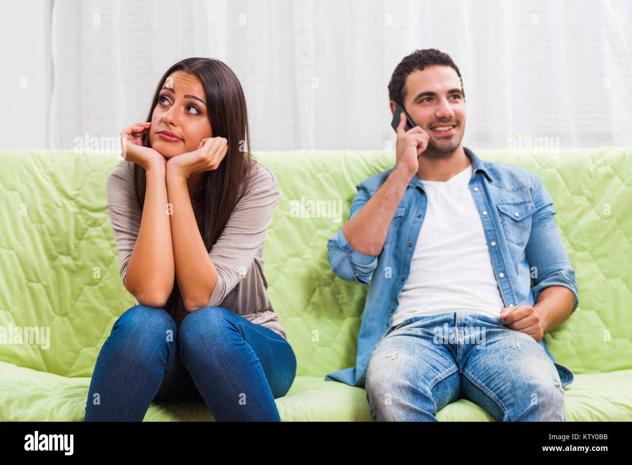 Young couple is sitting on sofa at home. Woman is getting bored while man is typing on phone. Stock Photo