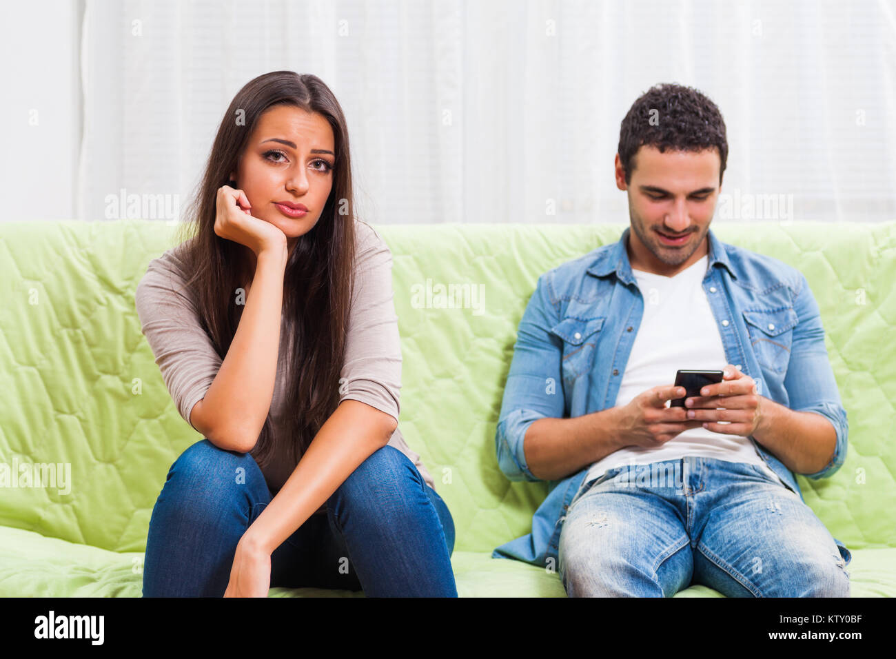 Young couple is sitting on sofa at home. Woman is getting bored while man is typing on phone. Stock Photo