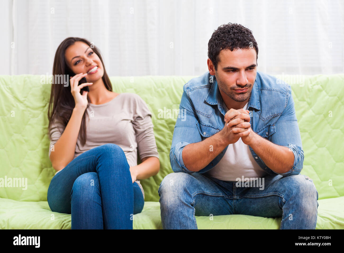 Young couple is sitting on sofa at home. Man is getting bored while woman is talking on phone. Stock Photo